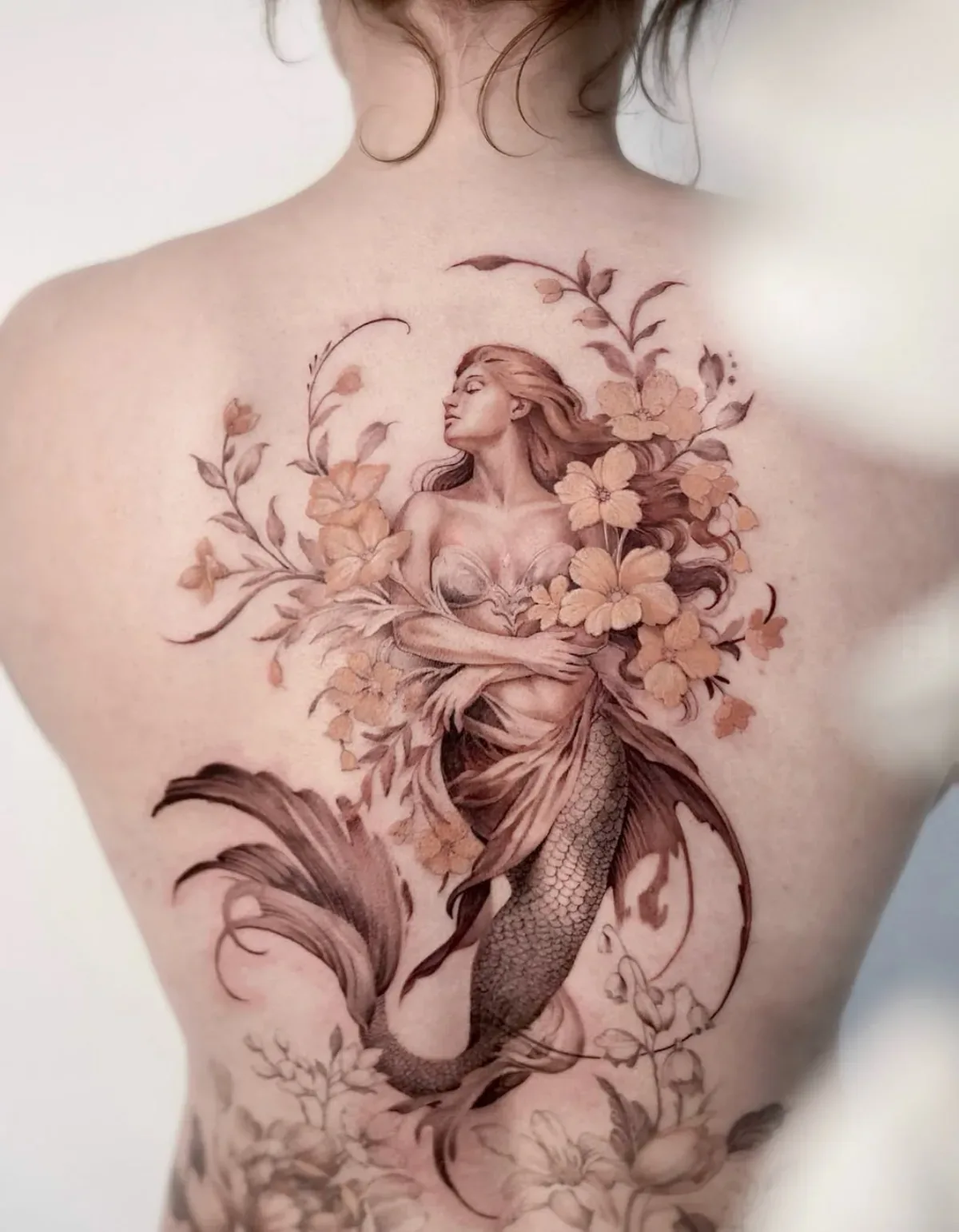 Meerjungfrau zwischen Blüten und Ranken auf dem Rücken. Fine-Line-Tattoo in Black & Grey mit zarten Farbakzenten von Annlie.